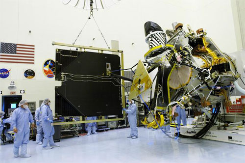 In this image, workers wearing blue coveralls (called 'bunny suits'), white hair bonnets and face masks work on the immense Mars Reconnaissance Orbiter in a large, white cleanroom at NASA's  Kennedy Space Center.  On the left side of the image the technicians and engineers are removing one of the large solar panels, about the size of a billboard.  On the right side of the image is the spacecraft itself, a large portion of which is covered in protective gold and black thermal blanketing.  Multiple cords, used for power and testing, hang from the massive spacecraft bus.