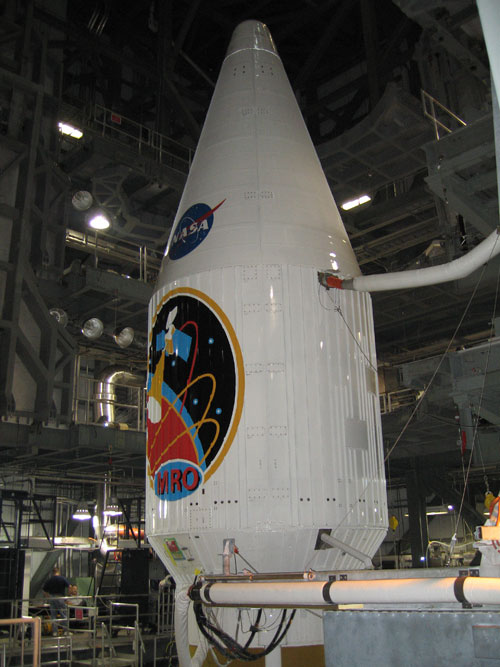 In this image, in the center, is the large white, protective fairing bearing both the red, white and blue NASA 'meatball' logo and the very colorful mission logo.  The fairing sits atop the large Atlas V rocket that will launch it out of Earth's atmosphere, toward Mars.  The inside of the vertical integration facility is very industrial looking with large steel beams, tubing and service stairways.  There is a tube attached to the fairing that provides air conditioning to the spacecraft.