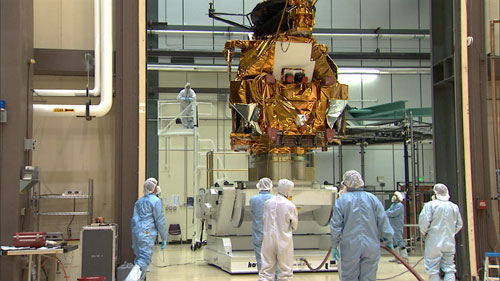 A group of eight engineers and technicians dressed in white and blue cleanroom coveralls and bonnets (called 'bunny suits') stand and watch as the large, boxy bus of the Mars Reconnaissance Orbiter is lifted by a large, white mechanism.  The bus is covered in protective, gold thermal blanketing.