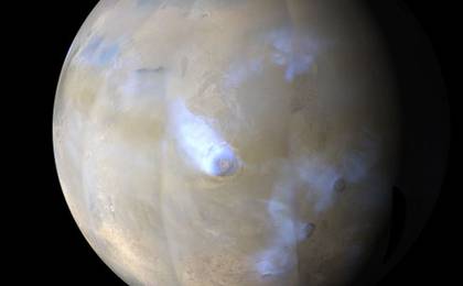 This image shows a simulated view of Mars from orbit, looking at the planet from the north. White patches streaming from Olympus Mons and other Martian volcanoes are clouds of water ice.White patches at the top of the globe are north polar ice deposits. Hazy, brown streaks that obscure the surface are regional dust storms. The planet's surface appears gray-blue and tan.