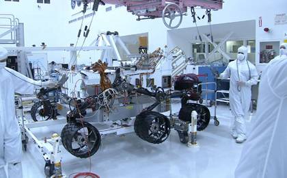 In this picture, the Curiosity rover is sitting on top of six shiny wheels.  The picture was taken from the side of the rover and only four wheels are visible.  The wheels have a black coating which makes them slip resistant.  On the right of the rover is an engineer who is wearing a white 'bunny suit' to prevent him from getting any unwanted Earth microbes onto the rover.  The engineer is holding one of the lift cables used to put the rover onto the wheels.  A pink heavy-lift crane is visible over the rover.  The crane is used to lift the rover body within the room.  Behind the rover, lots of lab equipment and other parts are scattered around the cleanroom.
