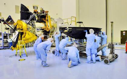 Technicians and engineers dressed from head to toe in blue coveralls (called 'bunny suits') prepare the Mars Reconnaissance Orbiter's large, black high-gain antenna (that resembles a satellite dish) to be mated with the body of the spacecraft.  The bus of the spacecraft with nearly all of its instruments in place, sits just to the left of the people pictured.  Parts of the orbiter are covered in gold, shiny thermal blanketing.  Facing directly upward is the spacecraft's huge Hi-RISE camera covered in black protective blanketing.