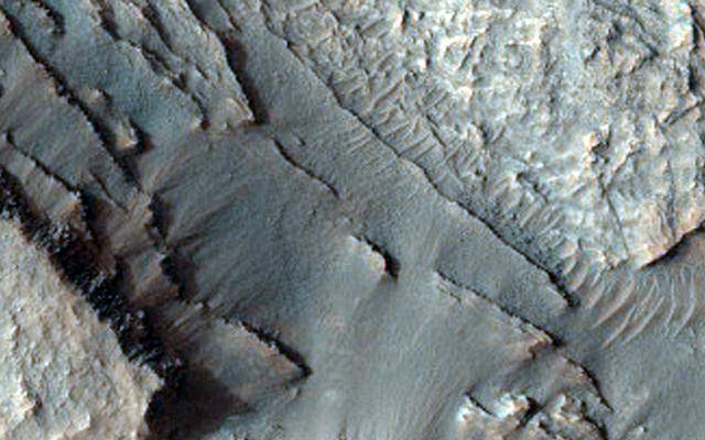 This image shows part of the floor of an impact crater on the northern rim of the giant Hellas Basin. 

Hellas includes the lowest elevations on Mars, and may have once held lakes or seas; layered rock outcrops occur around much of the edge of the basin. At this site, a large impact crater (about 90 kilometers across) was partly filled by layered rocks. These rocks on the crater floor are now eroding and forming strange pits.

Here, the layers are mostly exposed on a steep slope which cuts across much of the image. On this slope, they crop out as rocky stripes, some continuous and others not. The material between the stripes is mostly covered by debris, but some areas of exposed rock are visible. The slope is capped by a thick, continuous layer that armors it against erosion; once this cap is gone, the lower material is removed rapidly, forming the steep slope. At the base of this slope, rocks on the floor of the pit appear bright and heavily fragmented by cracks known as joints.

The variation in rock types suggests that the rocks here were deposited by multiple processes or in different environments. Sites like this may preserve a record of conditions on early Mars.