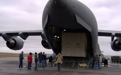 In this image, more than a dozen people stand, in winter coats, behind a huge, gray military C-17 cargo plane.  From this vantage point, the plane resembles a huge shark with its mouth wide open.  Inside the 'mouth' is one of the large boxes that contain the Mars Reconnaissance Orbiter.  The wings of the plane jut out from both sides.  The sky is cloud-covered.