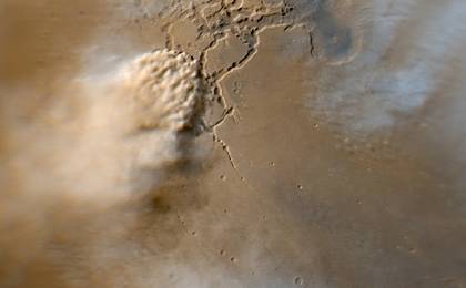 This image shows a turbulent mass of thick, roiling, red Martian dust rising from a network of canyons and flowing diagonally toward the lower left corner of the frame. Above the storm front, wispy, white clouds of water ice trail off toward the left, toward the bottom of the frame, toward the top of the frame, and toward the upper right corner of the frame.