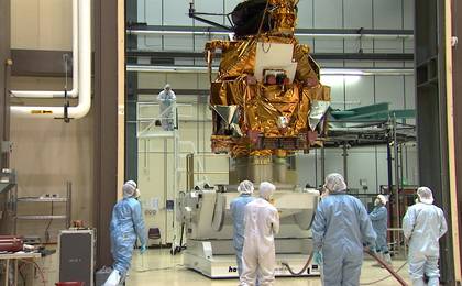 A group of eight engineers and technicians dressed in white and blue cleanroom coveralls and bonnets (called 'bunny suits') stand and watch as the large, boxy bus of the Mars Reconnaissance Orbiter is lifted by a large, white mechanism.  The bus is covered in protective, gold thermal blanketing.