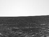 A black and white panoramic image showing layers of rocks on the right and left side of the image.  The rover's antenna is visible on the bottom-right and the solar panels can be seen on the bottom right.