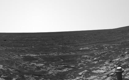 A black and white panoramic image showing layers of rocks on the right and left side of the image.  The rover's antenna is visible on the bottom-right and the solar panels can be seen on the bottom right.