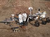 Two spacecraft engineers stand with a group of vehicles providing a comparison of three generations of Mars rovers developed at NASA's Jet Propulsion Laboratory, Pasadena, Calif. The setting is JPL's Mars Yard testing area.