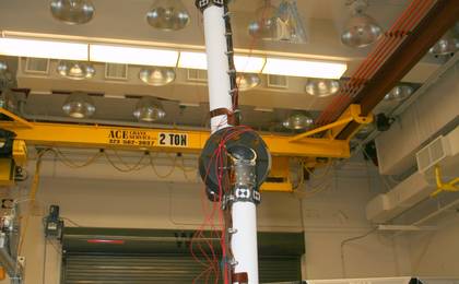 In the middle of this image, the long robotic arm rises straight up toward the ceiling of the lab where it is being tested.  The arm is white, and is flexible at several gray round 'joints.'  Red wires dangle and are strung along various parts of the arm.  At the end of the arm is a complicated set of instruments.  In the background, a yellow crane for lifting equipment up to 2 tons (4,000 pounds) can slide along the ceiling to place heavy equipment anywhere in the room.  A metal garage-like door and computer stations lie behind the area where the arm is extended.