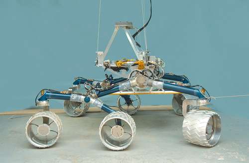 In this image, a large, six-wheeled rover sits on a concrete floor in a laboratory against a light-blue background.  The rover's 'legs,' or mobility system are a medium blue color and the large, cleated wheels are shiny silver.  On top of the mobility system is a silver-colored triangle that represents where the heart and brains of the rover will go when it is complete.