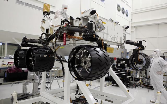 This image was taken in the clean room where the Curiosity rover is being assembled.  It shows the rover, which is about the size of an SUV, hoisted on a white lift, with its black wheels suspended in the air.   One engineer is on top of the hoist and is leaning over the rover body, while another is looking up on the ground floor to the right of the rover.  Both engineers are wearing white "bunny suits" to keep them from contaminating any equipment.