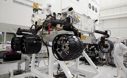 This image was taken in the clean room where the Curiosity rover is being assembled.  It shows the rover, which is about the size of an SUV, hoisted on a white lift, with its black wheels suspended in the air.   One engineer is on top of the hoist and is leaning over the rover body, while another is looking up on the ground floor to the right of the rover.  Both engineers are wearing white "bunny suits" to keep them from contaminating any equipment.