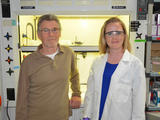 Two scientist are pictured are pictured in the icy worlds lab.  On the left is Michael Russell wearing a blown shirt and rimmed glasses.  Laurie Barge is on the right and she is wearing a lab coat and protective lab glasses.  In the background is a machine lit up by a glowing yellow light.