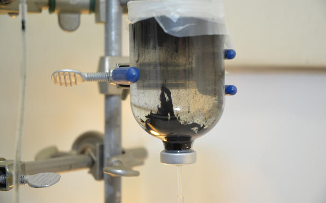 This image shows a glass bobble, upside down, like an IV hanging from a metal post. Through the clear glass there is a clear liquid with black "goo" accumulating toward the bottom of the bottle.