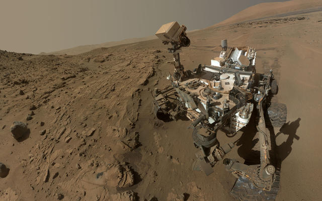Curiosity, rover, self-portrait, rock, drilling, windjana, waypoint, kimberley - This image is a self-portrait of the Curiosity rover taken from the site with the rover's 'head' down, with rock formations surrounding it and Mount Sharp in the background.