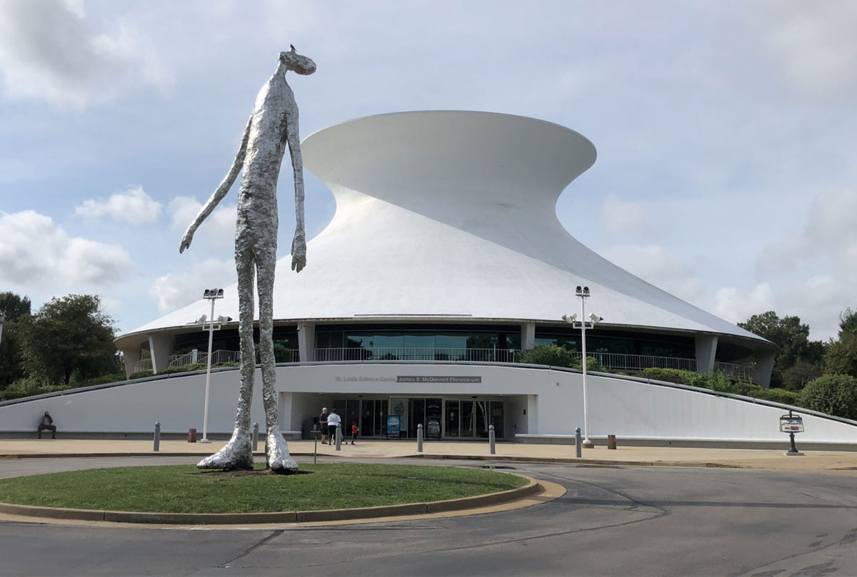 An image of a tan modern building with a dinosaur sculpture in front