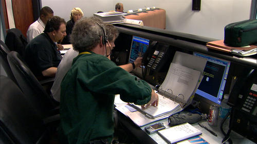 In this image, six individuals sit at consoles in front of computers during a launch rehearsal for the Mars Reconnaissance Orbiter mission. Launch vehicle manager Arden Acord (a gray-haired, Caucasian middle-aged man with a goatee) points to data on project manager Jim Graf's computer. Graf is a brown-haired Caucasian man in his fifties. Flight system manager Howard Eisen sits to Graf's left. He is a Caucasian man with brown hair and a goatee. Next to Eisen are two Lockheed Martin employees, one of whom is Tammy Harrington, the mission integration manager. She is a blond woman in her forties.