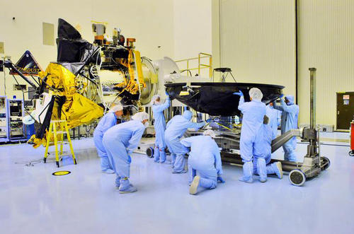 Technicians and engineers dressed from head to toe in blue coveralls (called 'bunny suits') prepare the Mars Reconnaissance Orbiter's large, black high-gain antenna (that resembles a satellite dish) to be mated with the body of the spacecraft. The bus of the spacecraft with nearly all of its instruments in place, sits just to the left of the people pictured. Parts of the orbiter are covered in gold, shiny thermal blanketing. Facing directly upward is the spacecraft's huge Hi-RISE camera covered in black protective blanketing.