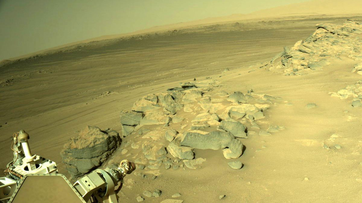 slide 3 - This is an image of the Mars landscape taken by the Perseverance rover. A clear sky, some hills and sandy ground are present int he horizon. Up close there are many large boulders and a part of Perseverance’s AutoNav camera and its shadow are present.