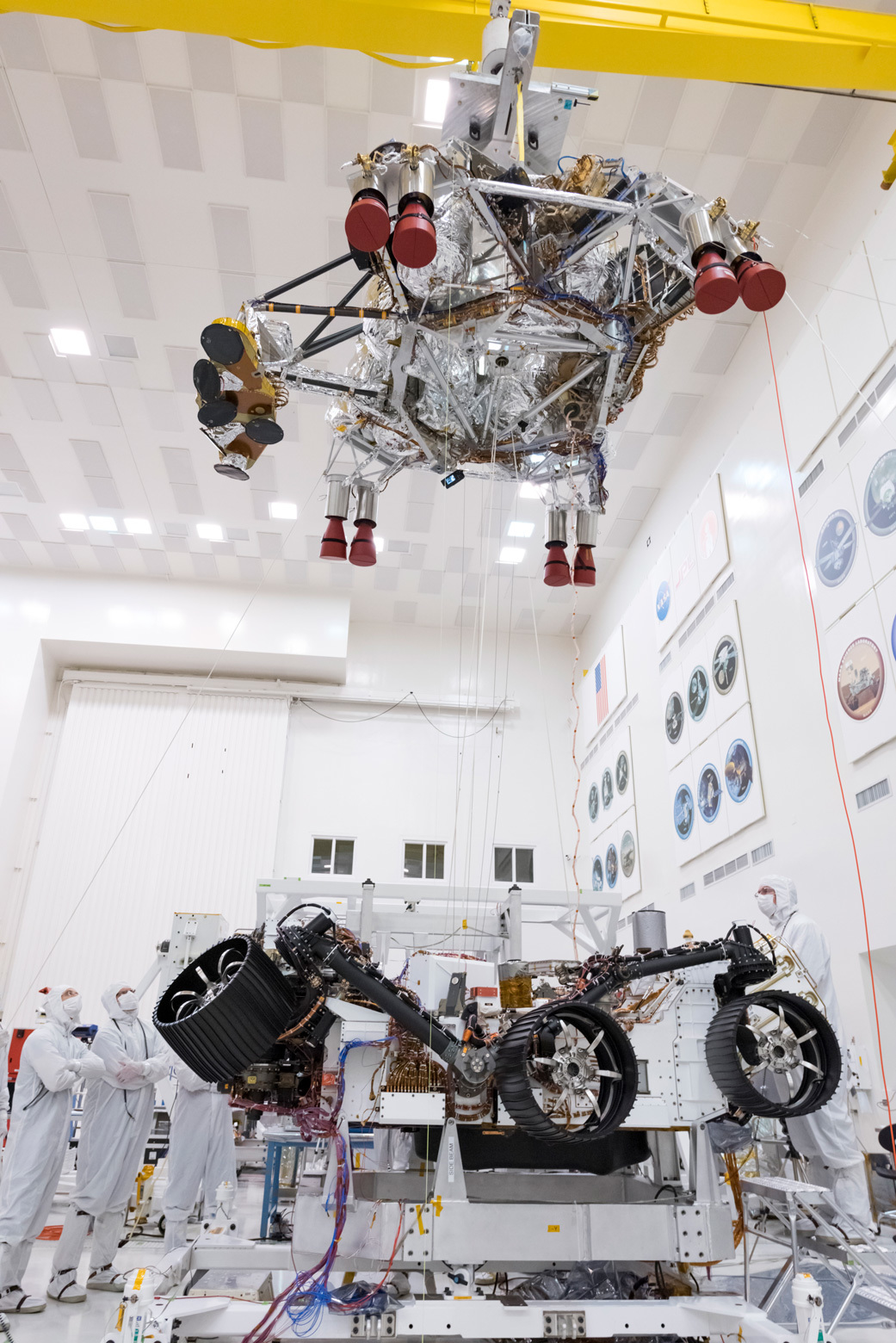 Engineers and technicians working on the Mars 2020 spacecraft, look on as a crane lifts the rocket-powered descent stage away from the rover after a test. 