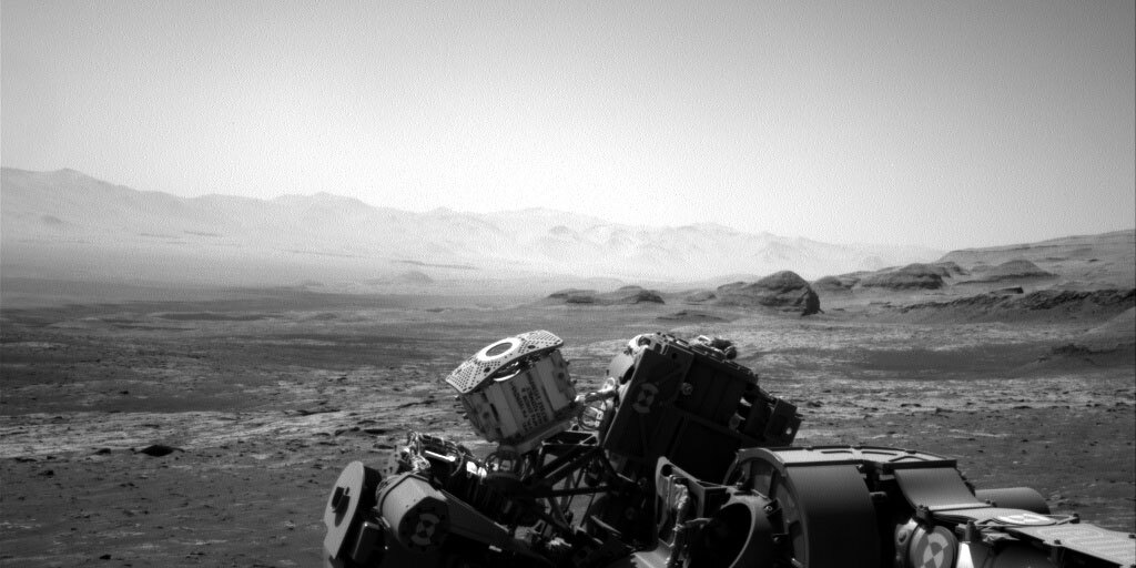 This is a black and white image of the sandy landscape of Mars. The front of the image displays a part of the Curiosity rover. About a handful of large rocks of different textures are to the left of the image. There are many hills dimly  displayed in the background. 