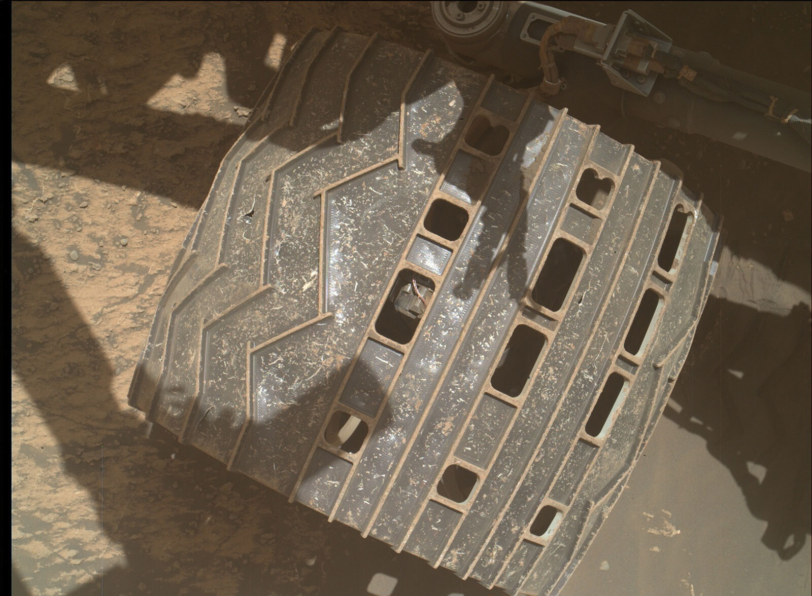 This is a large colored and zoomed in image of one of the wheels on Curiosity. The wheel is located on the sandy surface of Mars. Curiosity's shadow is reflected on the surface. 