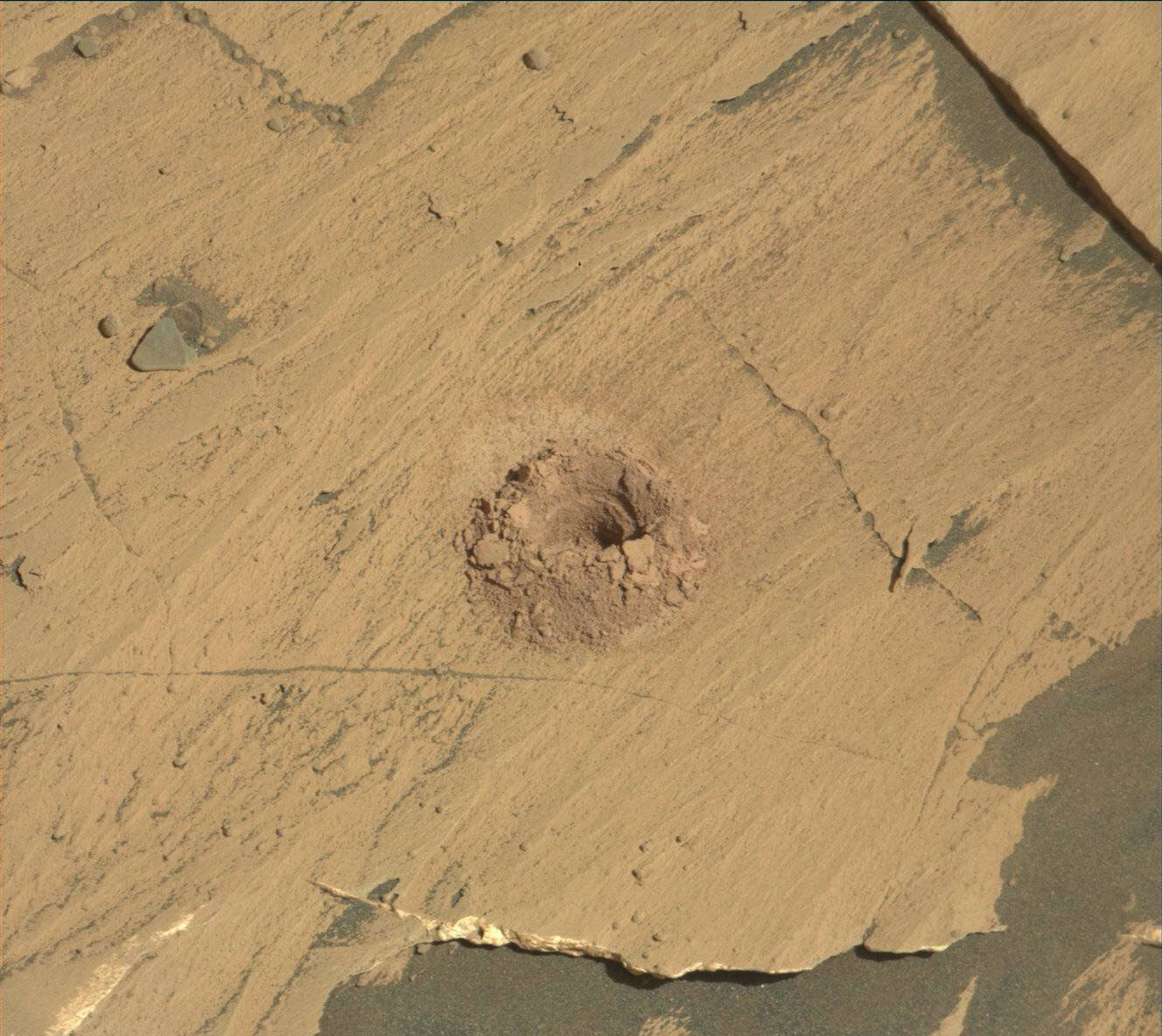 This is an image of the sandy surface of Mars. There are several cracks in the surface and a quarter sized hole with grainy crushed sand surrounding the hole. Curiosity’s shadow is in the bottom right corner. 