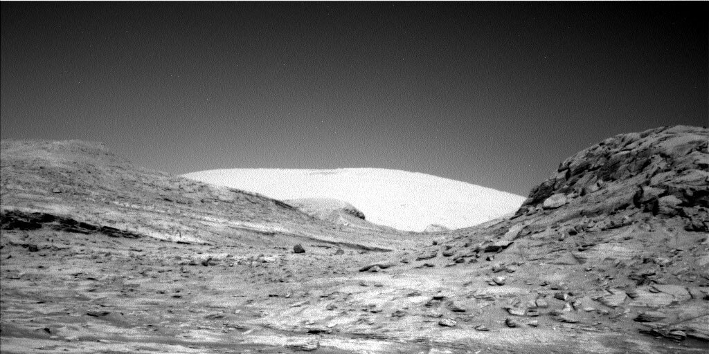 This is a black and white image of the Mars landscape taken by the Curiosity rover. A clear sky, some low, rocky hills and sandy ground are present in the horizon of the image.