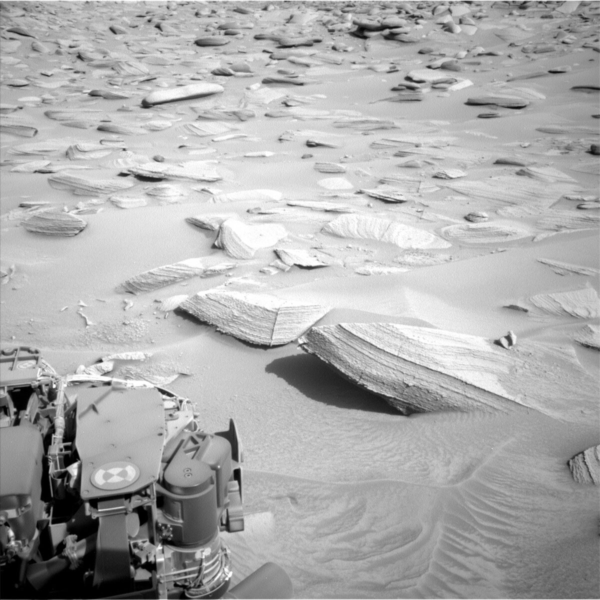 This image shows part of the Curiosity rover in the bottom left and rock formations on the Mars surface up ahead.