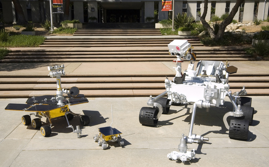 This image shows the front sides of three rovers viewed at an angle. On the right is the Mars Science Laboratory rover, which is the size of a small sport utility vehicle. Its 7-foot-long arm is extended in front of it, resting on the pavement. The camera 'eyes' at the top of its mast are turned downward at its smaller predecessors. In the middle, the tiny, wagon-size Sojourner rover has a deployable, alpha-particle X-ray spectrometer and single-color camera attached at its rear. On the left, the dune-buggy-size Mars Exploration Rover has its robotic arm extended in front of it, resting on the pavement. The panoramic camera 'eyes' at the top of its mast are turned upward, as if 'looking' at the bigger Mars Science Laboratory rover. All three rovers have six wheels, rocker-bogey suspension systems, rectangular bodies, solar panels, and antennas pointing upward from their spacecraft decks.
