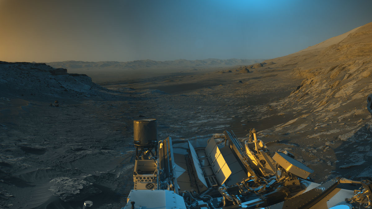 NASA’s Curiosity Mars rover used its black-and-white navigation cameras to capture panoramas of this scene at two times of day. Blue, orange, and green color was added to a combination of both panoramas for an artistic interpretation of the scene.