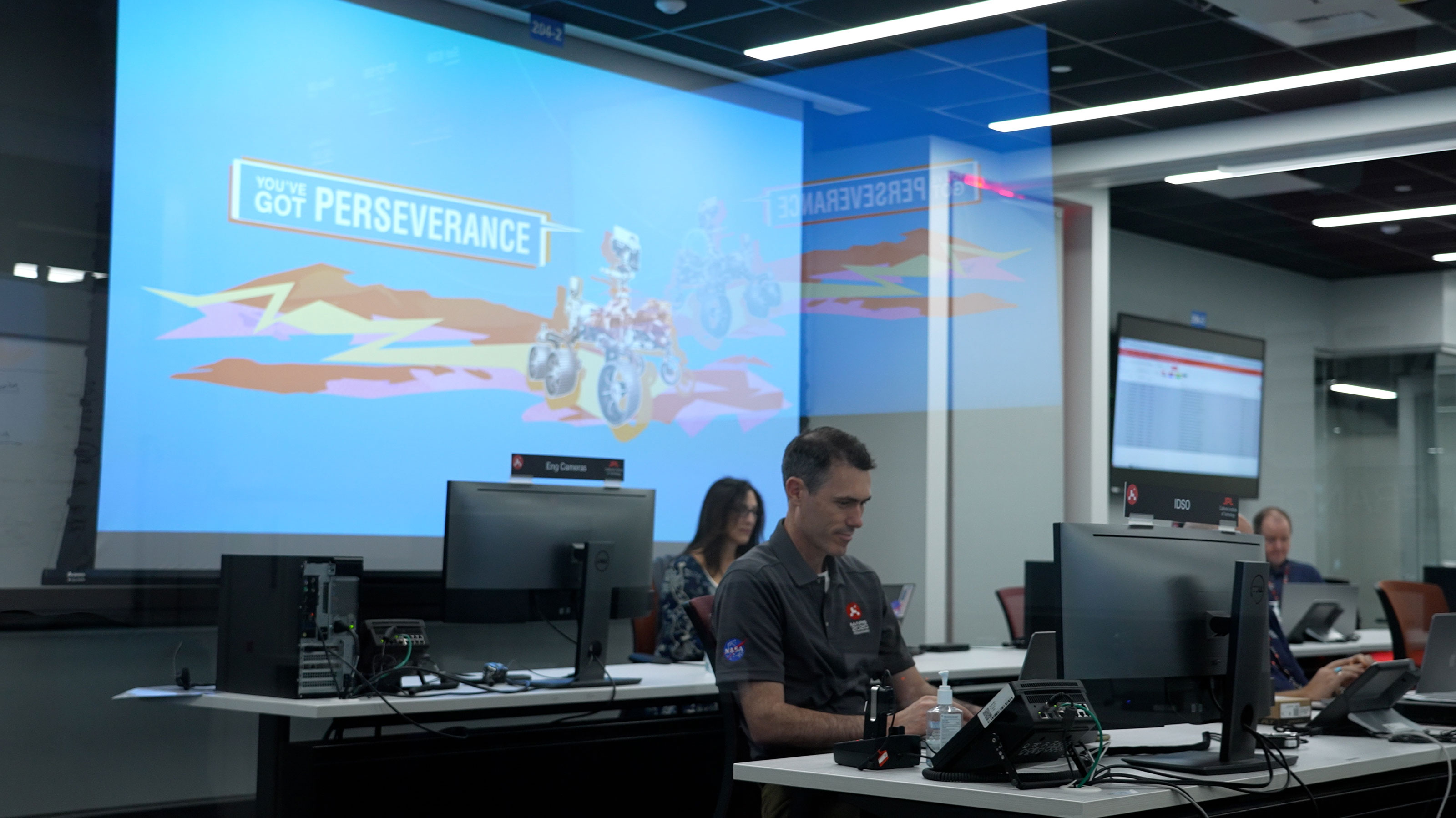 Image inside the control room for NASA's Perseverance rover shows the multi-colored "You've Got Perseverance" logo and illustration of the rover on one large screen on the wall, with two rows of tables and computers in front, with several Mars team members interacting virtually with student honorees.