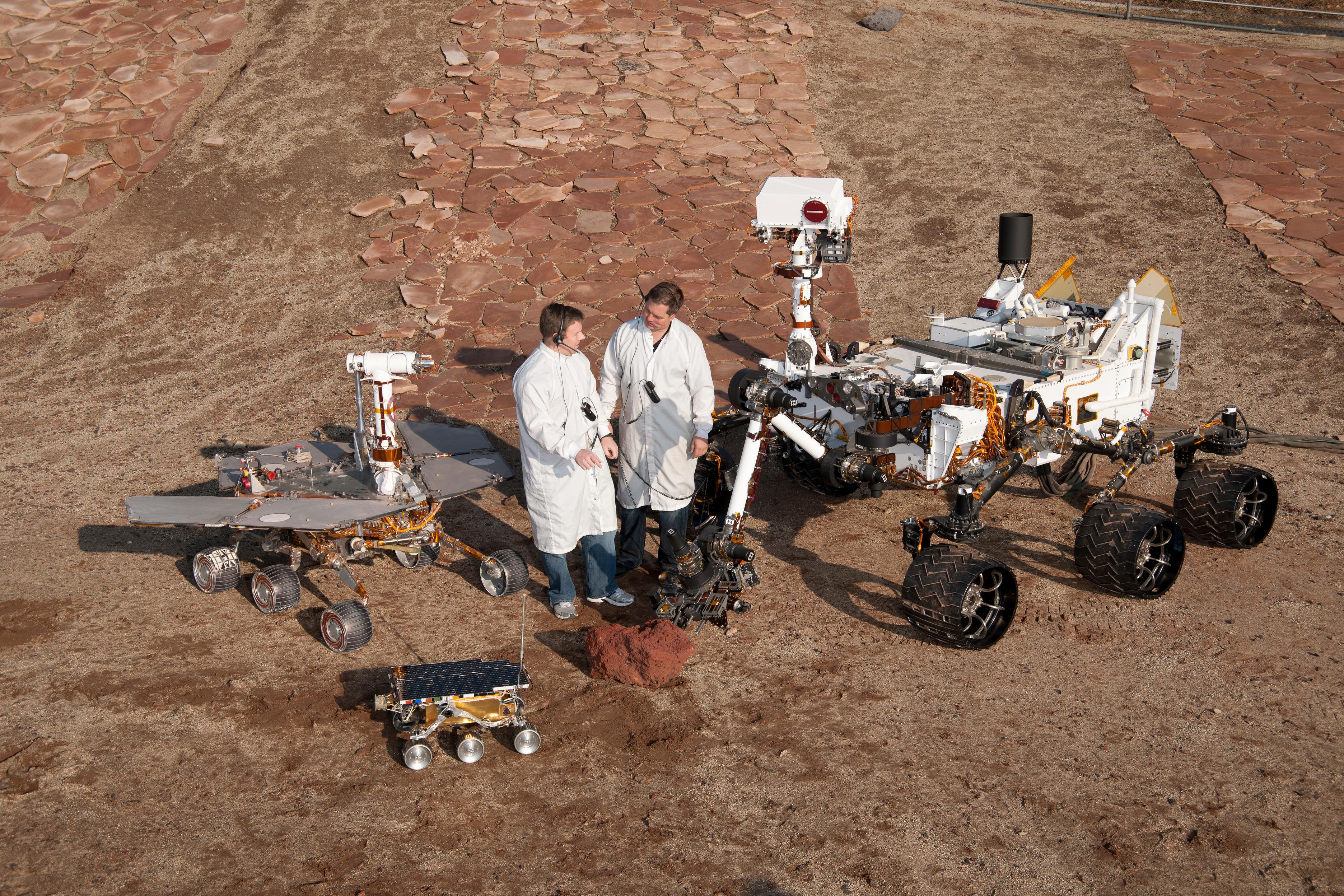Two spacecraft engineers stand with a group of vehicles providing a comparison of three generations of Mars rovers developed at NASA's Jet Propulsion Laboratory, Pasadena, Calif. The setting is JPL's Mars Yard testing area.