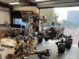 Engineering models of the Curiosity Mars rover (foreground) and the Perseverance Mars rover share space in the garage at JPL’s Mars Yard. 